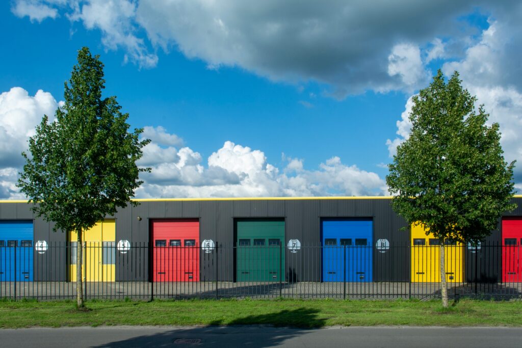 pexels photo 32038719 32038719 Vibrant garage doors in different colors with bright sky and clouds above, enclosed by iron gates.